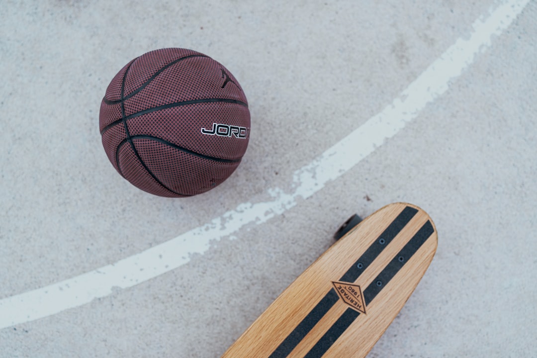 A maroon Jordan basketball and a wooden skateboard with 'HERITAGE 1960' branding rest on a light grey concrete court with...